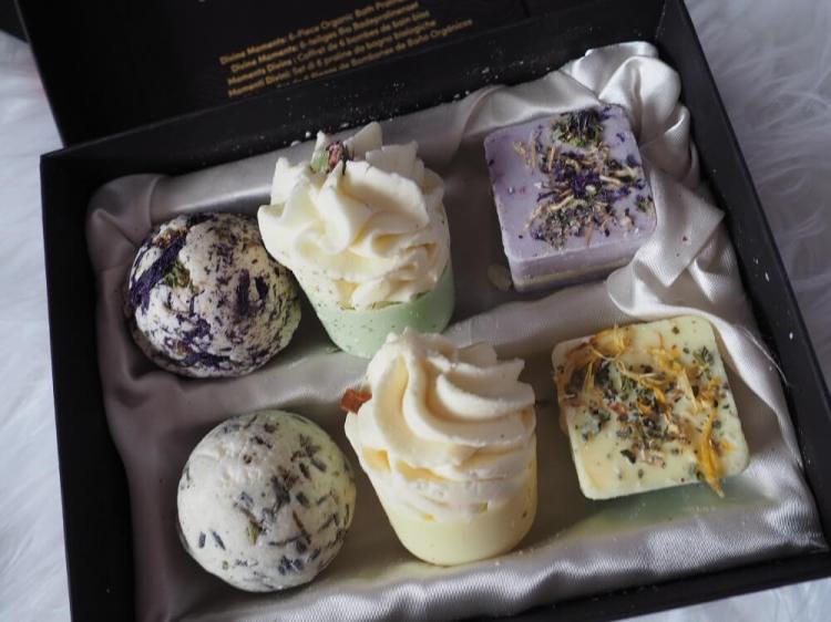 Geöffnete schwarze Geschenkbox mit goldener Prägung, sechs Satin Naturel Badepralinen, die beiden mittleren wie kleine Cupcakes mit Sahnehaube aussehen, auf edlem Satinstoff gebettet.