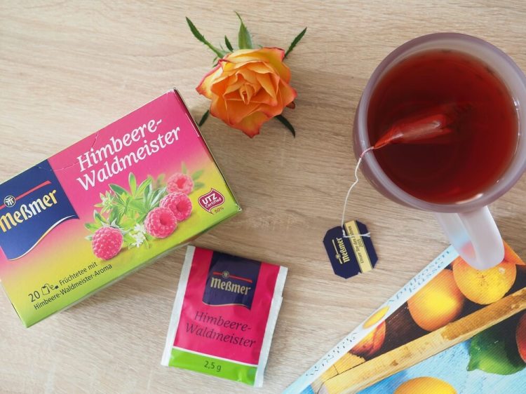Tasse mit Meßmer Himbeere-Waldmeister Tee, Teebeuteltütchen, Verpackung, dekoriert mit Buchecke und Rose