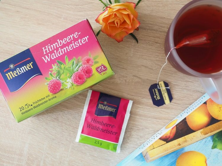 Meßmer Tee Himbeere-Waldmeister: Tasse mit Teebeutel, Teebeuteltütchen, Verpackung, Buchecke und Rose dekorativ arrangiert
