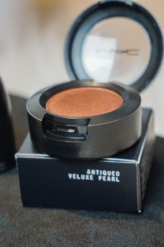 Nahaufnahme des MAC Single Eyeshadows „Antiqued Veluxe Pearl“ – eleganter Braunton mit seidigem Schimmer in der runden schwarzen Verpackung.