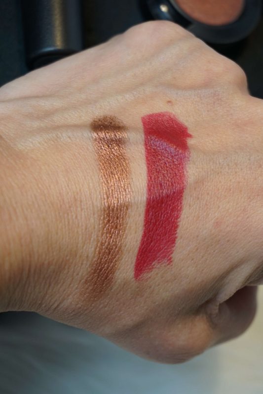 Swatches von MAC Eyeshadow „Antiqued Veluxe Pearl“ und MAC Lippenstift „D for Danger“ auf dem Handrücken – schimmerndes Bronze und sattes Beerigrot im direkten Vergleich.