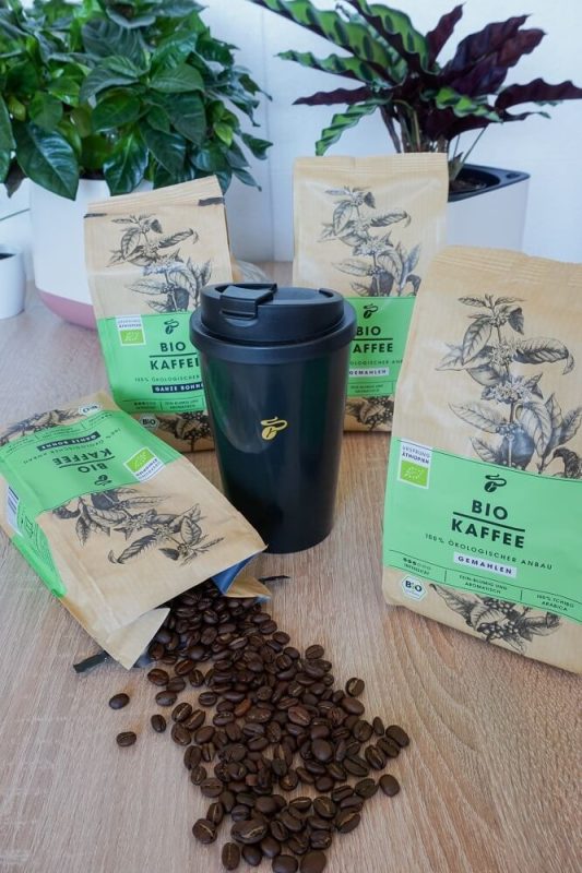 Schwarzer Tchibo Coffee-to-go-Becher im Vordergrund, daneben drei Packungen Bio Kaffee. Eine geöffnete Packung liegt neben dem Becher, Kaffeebohnen sind ausgestreut – nachhaltiger Kaffeegenuss im Test.