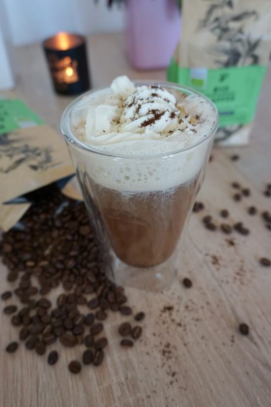 Glas mit frisch aufgebrühtem Kaffee und Sahnehaube im Vordergrund, daneben eine Tchibo Bio Kaffee Packung mit ausgestreuten Kaffeebohnen – Genussmoment pur.