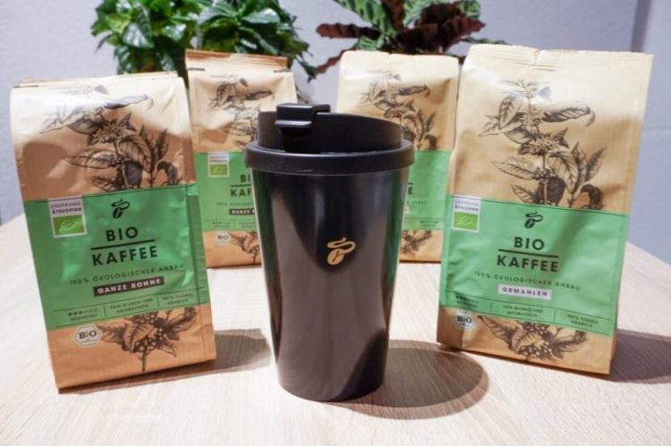 Schwarzer Tchibo Coffee-to-go-Becher mit Logo im Vordergrund, dahinter vier Packungen Tchibo Bio Kaffee – nachhaltiger Kaffeegenuss im Test.