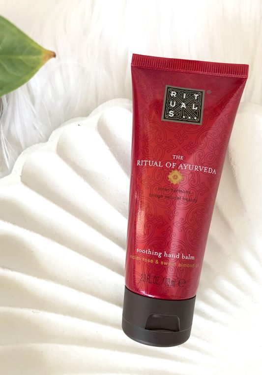 Rituals Handcreme The Ritual of Ayurveda liegt in Muschelschale aus weißem Holz auf weißem Fell – luxuriöses Pflege-Setting.