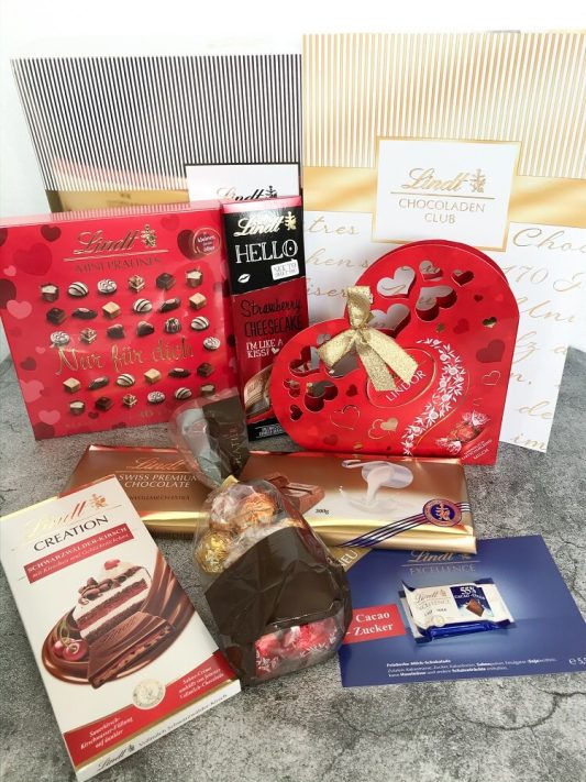 Hochkantaufnahme der Lindt Schokoladen-Club Box Februar 2020 „Valentin“ – alle enthaltenen Schokoladen und Pralinen dekorativ aufgereiht.