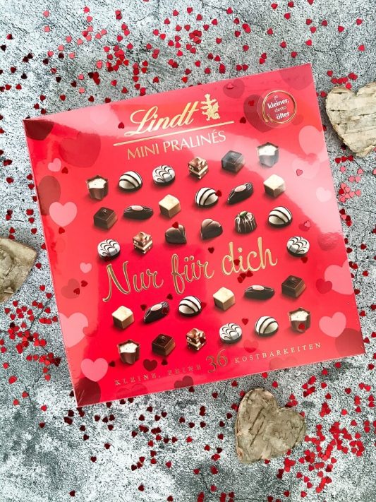 Verpackung der Lindt Mini-Pralines „Nur für dich“ auf Tisch dekoriert, umgeben von kleinen Herzchen-Pailletten – liebevoll inszeniert.