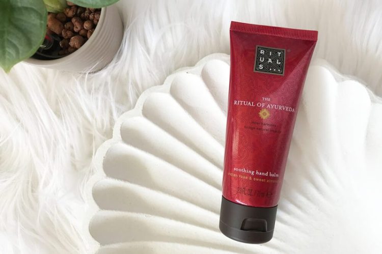 Rituals Handcreme The Ritual of Ayurveda in einer dekorativen Muschelschale aus weißem Holz, platziert auf weichem, weißem Fell.